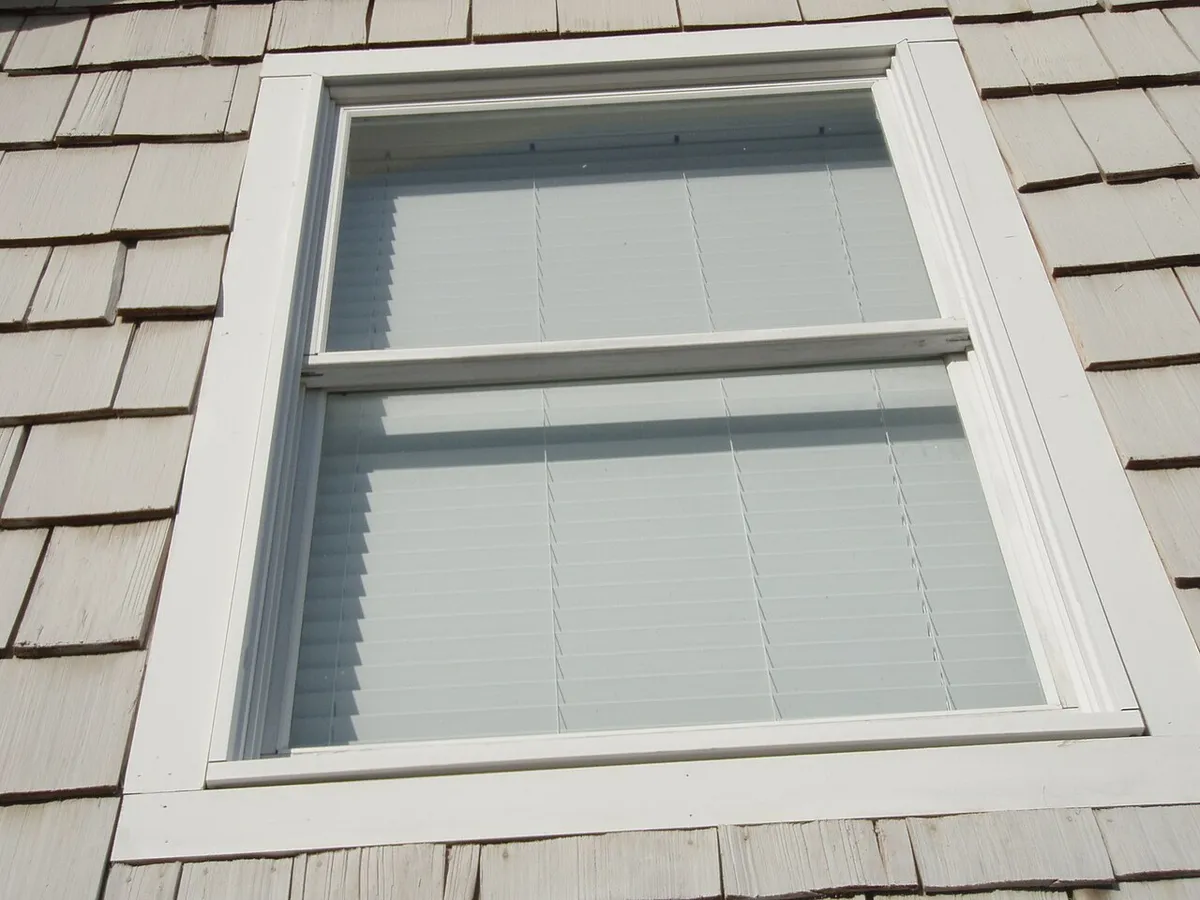A double-hung window with the blinds down, set in a shingled wall
