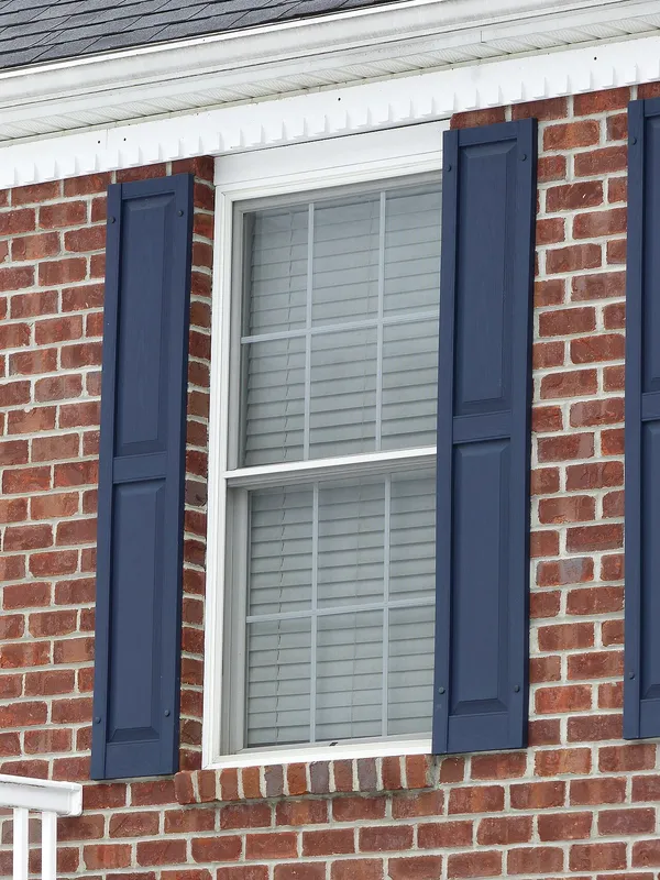 A white double-hung window with grids in a red brick wall, flanked by blue shutters