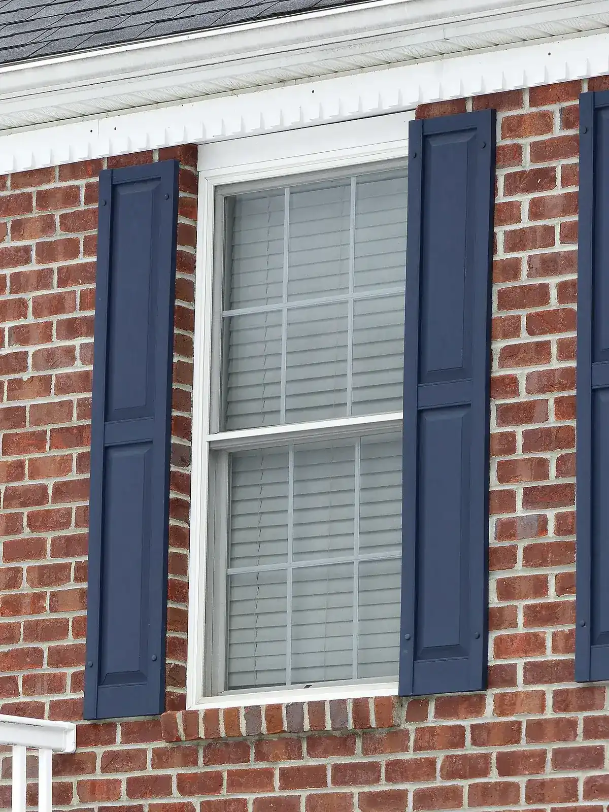 A white double-hung window with grids in a red brick wall between blue shutters