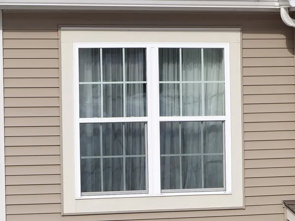A white double-hung window with grids and curtains in a tan vinyl-sided wall