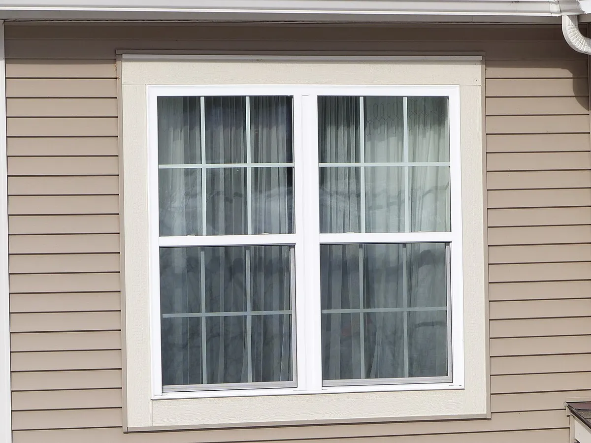A white double-hung window with grids and curtains in a tan vinyl-sided wall